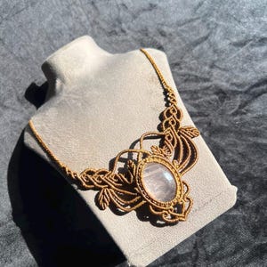 Puede incluir: Un collar de macramé con una piedra preciosa central de color rosa claro translúcido y forma ovalada. La piedra está rodeada por un intrincado trabajo de cordón marrón anudado que se extiende en un patrón decorativo con formas de hojas.
