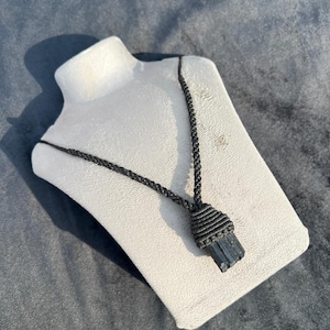 May include: A black tourmaline pendant necklace. The necklace features a raw, rectangular black tourmaline crystal wrapped in a macrame design. The cord is black and the pendant is displayed on a white bust.