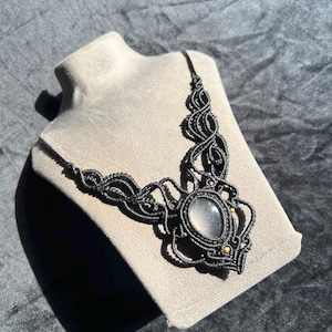 Puede incluir: Un collar de macramé negro con una piedra central translúcida en forma de lágrima. El intrincado diseño de cordones anudados se extiende desde la piedra, acentuado con pequeñas cuentas redondas de color dorado incrustadas en el patrón oscuro.