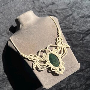 Puede incluir: Un collar de macramé con una piedra de color verde oscuro en forma ovalada en el centro. La piedra está rodeada por intrincados patrones de cordón anudado en color blanco roto que se extienden hacia afuera para formar la cadena del collar.