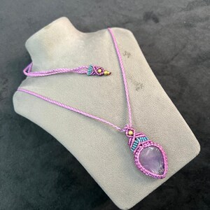 Op de afbeelding: Handgemaakte macramé ketting met een grote, hartvormige amethist hanger. De ketting heeft een lavendelkleurig koord met turquoise en gouden accenten. De hanger is dieppaars van kleur en wordt getoond op een grijze mannequin.
