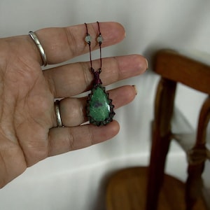 May include: A handcrafted pendant necklace featuring a teardrop-shaped green stone with red inclusions, intricately woven with dark brown cord. The necklace has a dark brown cord and small beads. The pendant is held in a hand.
