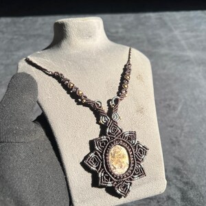 May include: A handmade macrame necklace with a starburst pendant and an oval stone at its centre. The necklace is made with brown and beige cords, and small gold-coloured beads. The pendant is displayed on a grey mannequin.
