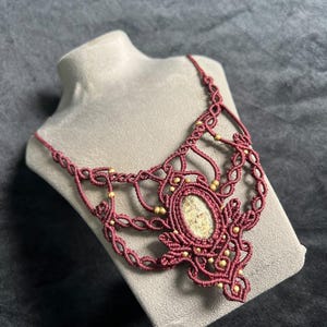 Op de afbeelding: Een macramé ketting met een centrale ovaalvormige steen met een gevlekt beige en bruin patroon, omringd door ingewikkeld donkerrood geknoopt koordwerk en kleine goudkleurige kralen die in het gevlochten ontwerp zijn verwerkt.