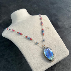 May include: A handcrafted necklace featuring a large, pear-shaped, iridescent blue gemstone pendant. The pendant is encased in a silver-toned wire frame. The necklace chain is strung with small red, blue, and clear beads, creating a colorful and elegant design.