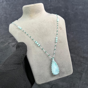 Op de afbeelding: Een delicate ketting met een druppelvormige hanger in lichtblauw en wit. De hanger hangt aan een zilveren ketting versierd met kleine groene en lichtblauwe kralen. De ketting wordt getoond op een grijze mannequin.