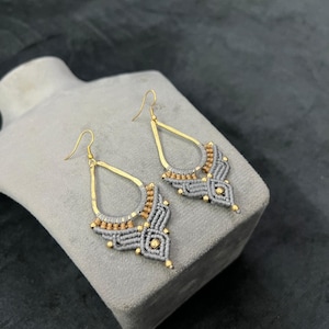 以下が含まれることがあります： ゴールドカラーの金属アクセントが付いた、手作りのグレーのマクラメイヤリングのペア。 イヤリングは、複雑な結び目と小さなゴールドビーズを備えたティアドロップ型です。 イヤリングは灰色の表面に表示されます。