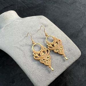 Puede incluir: Un par de pendientes de macramé hechos a mano. Los pendientes presentan un diseño tejido en color beige con detalles dorados y ganchos para las orejas dorados. Los pendientes se muestran sobre una superficie gris, sobre un fondo oscuro.