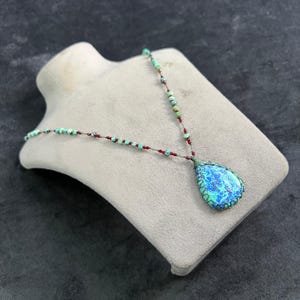 Puede incluir: Un collar artesanal con un colgante en forma de lágrima. El colgante tiene una piedra azul y verde, un borde verde y está suspendido de una cadena de cuentas. La cadena presenta cuentas turquesas y rojas. El collar se exhibe en un maniquí gris.