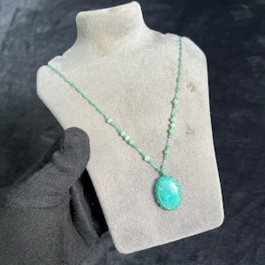 Può includere: Una collana verde con un grande pendente ovale. Il pendente è di colore turchese chiaro ed è circondato da una delicata cornice intrecciata. La catena della collana è fatta di piccole perline e filo verdi, esposta su un busto di gioielli grigio.