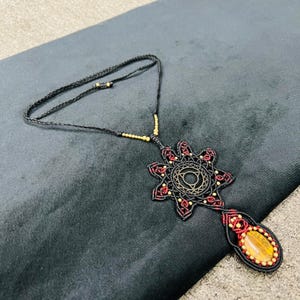 May include: A black macrame necklace with a starburst pendant featuring red accents and a central circular design. The pendant is connected to a braided black cord with gold-colored beads. A teardrop-shaped stone is at the bottom of the pendant.