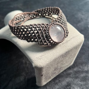 Puede incluir: Una pulsera de macramé hecha de cordón marrón oscuro con un complejo patrón de celosía tejida. Una piedra preciosa de cuarzo rosa, lisa, redonda y de color rosa pálido, está engastada en el centro de la banda, sujeta por el diseño anudado.