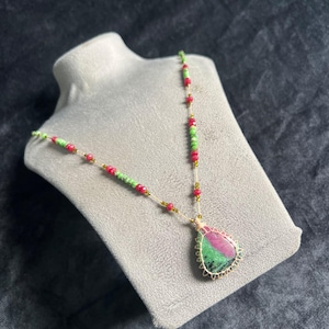 Puede incluir: Un collar con un colgante de rubí zoisita en forma de lágrima envuelto en alambre dorado. La cadena consiste en un cordón de color claro intercalado con pequeñas cuentas facetadas rojas y verdes dispuestas en un patrón repetitivo a lo largo.
