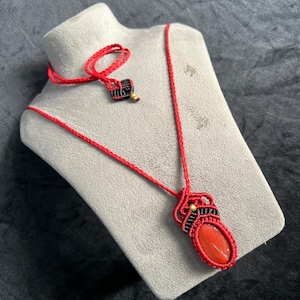 Könnte beinhalten: Eine rote Makramee-Halskette mit einem ovalen, rot-orangen Steinanhänger. Die Halskette wird auf einer grauen Büste präsentiert. Die Kordel ist rot, mit schwarzen und goldenen Akzenten in der Nähe des Anhängers. Handgefertigt.
