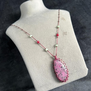 May include: A handcrafted necklace with a large, oval pink and black stone pendant, framed in reddish-brown woven material. The necklace has a matching reddish-brown cord, adorned with red and green beads. A unique piece of jewellery.