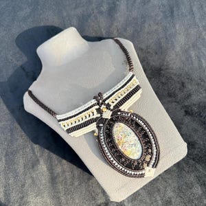 May include: A handmade macrame necklace with a large oval pendant. The pendant features a colorful, abstract design within a dark brown frame. The necklace has a brown cord and a white and gold accent design.