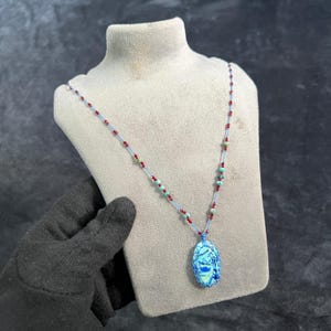 Puede incluir: Un collar con un colgante azul con un diseño blanco, ensartado en un cordón azul con cuentas rojas y turquesas. El collar se exhibe en un maniquí gris. El colgante tiene forma ovalada.