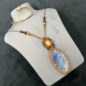 May include: A handcrafted necklace and bracelet set. The necklace features a large, oval blue stone pendant with a macrame design. The necklace and bracelet are made with brown and gold-colored cord and beads. The jewelry is displayed on a gray mannequin.