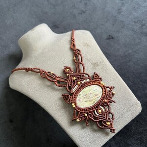 Può includere: Una collana macramè fatta a mano con un grande pendente ovale in pietra iridescente. La collana presenta un'intricata annodatura in un ricco colore marrone, impreziosita da piccole perline dorate. Il pendente è incastonato in una cornice macramè dettagliata.