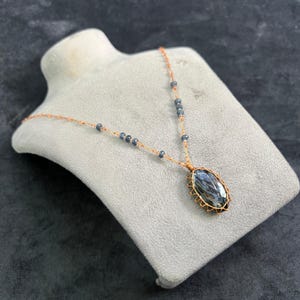 Puede incluir: Un collar artesanal con un gran colgante ovalado de piedra preciosa azul en un marco de alambre dorado. El collar tiene una delicada cadena dorada con pequeñas cuentas azules. El colgante se exhibe en un busto de joyería gris. Longitud: aprox. 45 cm.