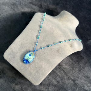 Puede incluir: Un collar artesanal con un colgante ovalado azul y verde. El colgante está engastado en un marco tejido azul y cuelga de una delicada cadena de cuentas azules. El collar se exhibe en un busto de maniquí gris.