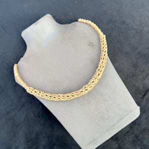 May include: A gold-colored necklace with a woven design is displayed on a gray mannequin. The necklace is a choker style, with a textured, braided appearance. The background is a dark, solid color.