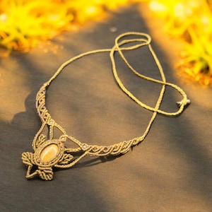 May include: A handcrafted gold-colored macrame necklace with an oval-shaped stone pendant. The necklace features intricate knotting and a delicate chain. The pendant is surrounded by a decorative macrame design. The necklace is displayed on a dark surface.