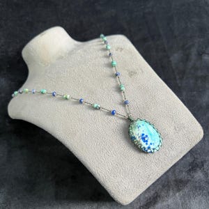 Puede incluir: Un collar artesanal con un gran colgante ovalado en azul claro y turquesa. El colgante está adornado con cuentas azules oscuras y verdes. El collar se exhibe en un busto de joyería gris.
