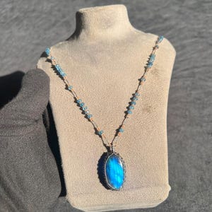 May include: A necklace featuring an oval labradorite pendant with a vibrant blue flash, encased in a silver wire-wrapped setting. The chain consists of small, light blue faceted beads interspersed with thin, metallic copper-coloured wire segments.