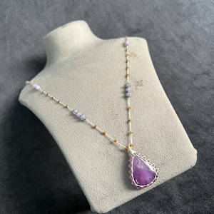 May include: A handmade necklace with a large, teardrop-shaped amethyst pendant. The pendant is set in a silver wire frame and hangs from a delicate chain with small gold and purple beads. The necklace is displayed on a grey mannequin.