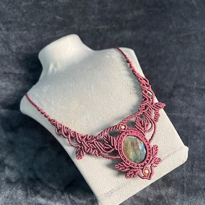 Può includere: Una collana in macramè rosso scuro con una pietra di labradorite ovale al centro. Il cordoncino è annodato in modo complesso con motivi a forma di foglia e piccole perline color oro integrate, creando un gioiello tessile molto dettagliato.