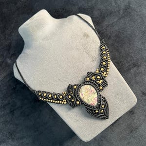 Op de afbeelding: Een zwarte macramé ketting met goudkleurige kralen en een centrale ovale steen met roze en groene tinten. De ketting wordt gepresenteerd op een grijze mannequin buste tegen een donkere achtergrond.