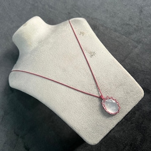 Puede incluir: Un delicado collar con un colgante de piedra preciosa ovalada y transparente. El colgante está encerrado en un marco de alambre y suspendido de un fino cordón rojo. El collar se exhibe sobre un busto gris, sobre un fondo oscuro.