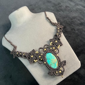 Peut inclure: Un collier en macramé fait à la main avec une grande pierre ovale turquoise. Le collier est fait avec un cordon marron foncé et des perles dorées. Le design est présenté sur un buste gris. Longueur d'environ 40 cm.