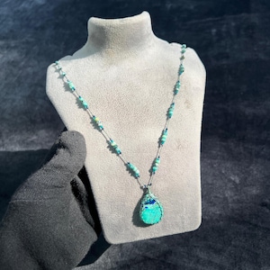 Puede incluir: Collar artesanal con colgante en forma de lágrima en tonos turquesa y azul. El collar está ensartado con pequeñas cuentas turquesas y un hilo azul oscuro, exhibido en un maniquí gris.