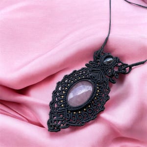 Puede incluir: Un collar de macramé negro con una gran piedra ovalada de cuarzo rosa como punto focal. El intrincado diseño presenta pequeñas cuentas doradas y una piedra más pequeña en la parte superior. El collar se muestra sobre un fondo de tela rosa.