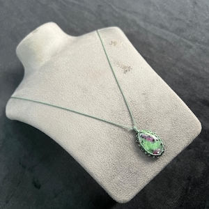 May include: A green cord necklace with an oval pendant. The pendant has a green and red stone, set in a silver-coloured frame. The necklace is displayed on a grey bust.