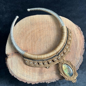 Op de afbeelding: Een handgemaakte ketting met een zilverkleurige kraag en een hanger met een iriserende steen. De ketting heeft een geweven, bruinachtige ontwerp met zwarte accenten en kleine zilveren kralen, liggend op een houten oppervlak.