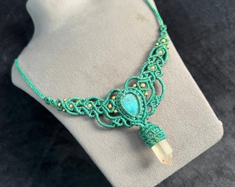 May include: A green macrame necklace with a turquoise stone and a clear quartz crystal pendant. The necklace is decorated with small gold beads. The necklace is displayed on a gray bust.