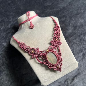 May include: A macramé necklace featuring a central oval-shaped stone with light green and grey tones, encased in dark rose-coloured knotted cord. The design includes intricate woven patterns, small gold-toned beads, and an adjustable cord closure.
