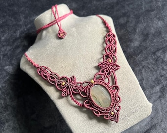 Puede incluir: Un collar de macramé con una piedra central de forma ovalada en tonos verde claro y gris, engastada en cordón anudado de color rosa oscuro. El diseño incluye intrincados patrones trenzados, pequeñas cuentas doradas y un cierre ajustable.