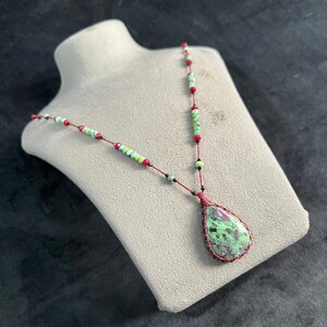 Op de afbeelding: Een handgemaakte ketting met een druppelvormige hanger met groene en rode steen. De ketting heeft een rood koord met groene, rode en zwarte kralen. De hanger is ongeveer 3,8 cm lang.