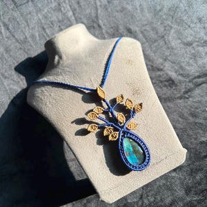 Op de afbeelding: Een macramé ketting met een druppelvormige labradoriet edelsteen met een blauwe glans, omvat door donkerblauw koord. Boven de steen is een boomachtig ontwerp gemaakt van beige koord, dat takken vormt met kleine blaadjes in knoopvorm.