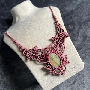Op de afbeelding: Een macramé ketting met een centrale ovale labradoriet edelsteen in iriserende blauwe en gouden tinten. De steen is gevat in donkerrood koord geweven in ingewikkelde bladvormige patronen, geaccentueerd met twee kleine goudkleurige metalen kralen.