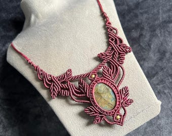 Pode incluir: Um colar de macramé com uma pedra central oval de labradorite em tons iridescentes de azul e dourado. A pedra está envolvida num cordão vermelho escuro tecido em padrões complexos de folhas, acentuado com duas pequenas contas metálicas douradas.