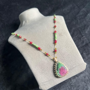 Puede incluir: Un collar con un colgante en forma de lágrima que presenta una piedra verde y magenta envuelta en alambre de color dorado. La cadena consiste en pequeñas cuentas verdes y rojas alternadas en un cordón fino y claro, creando un diseño delicado.