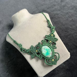 Puede incluir: Collar de macramé verde hecho a mano, con una piedra ovalada verde y azul. El collar se exhibe sobre un busto de maniquí. El diseño de macramé está adornado con pequeñas cuentas doradas. Fondo gris oscuro.