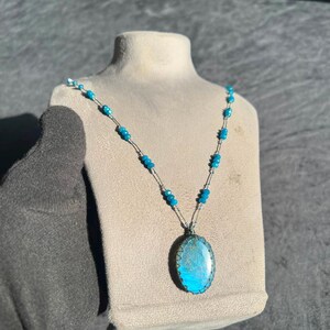 May include: A necklace featuring a central oval pendant with a blue iridescent stone set in a dark metal frame. The chain consists of small, faceted bright blue beads interspersed with thin silver-toned metal links, creating a delicate design.