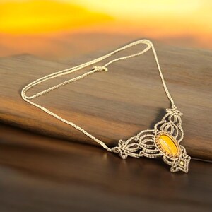 May include: A handcrafted macrame necklace with a central oval-shaped yellow stone. The necklace features intricate knotwork in a light beige color, with a delicate chain. The background is a warm gradient of orange and brown.