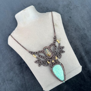 May include: A handcrafted macrame necklace featuring a turquoise gemstone pendant. The necklace has a dark brown cord with intricate knotting and leaf-like designs. Accents include small gold-colored beads and a tiger's eye stone.